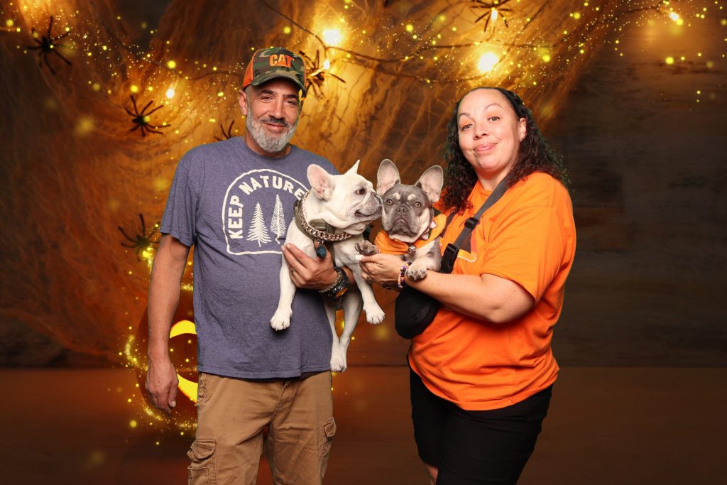Man and woman pose with their dogs taking a photo in front of a greenscreen Halloween Photo Booth.