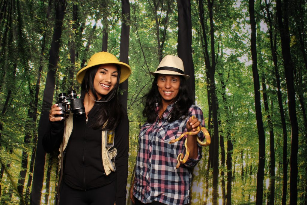 Two women in front of the forest backdrop posing for a picture in front of a SnapFiesta photo booth wearing camping themed props