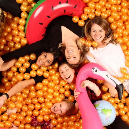 a group of women in an orange ballpit photobooth with inflatable toys and a watermelon innertube