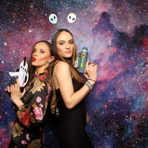 two women holding fake sci-fi weapons posing against a space background